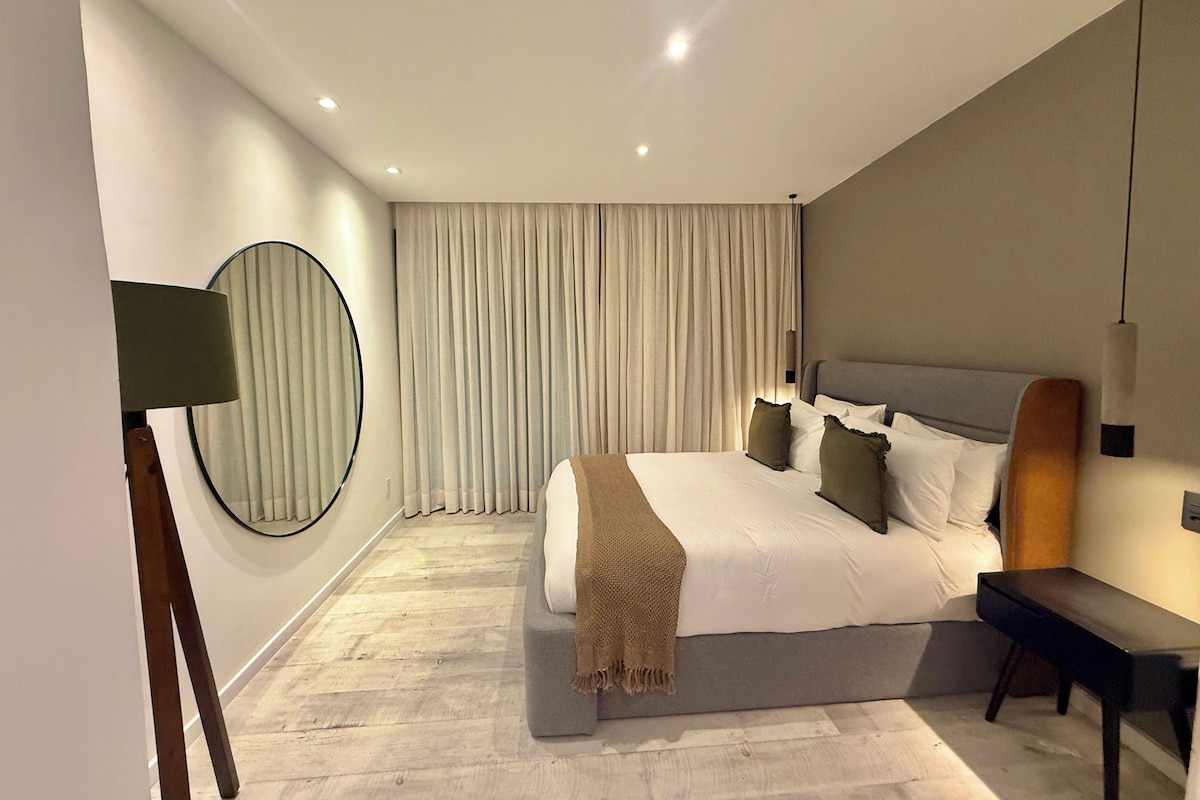 A modern bedroom with a large bed, a bedside table, a mirror, and a window with curtains.