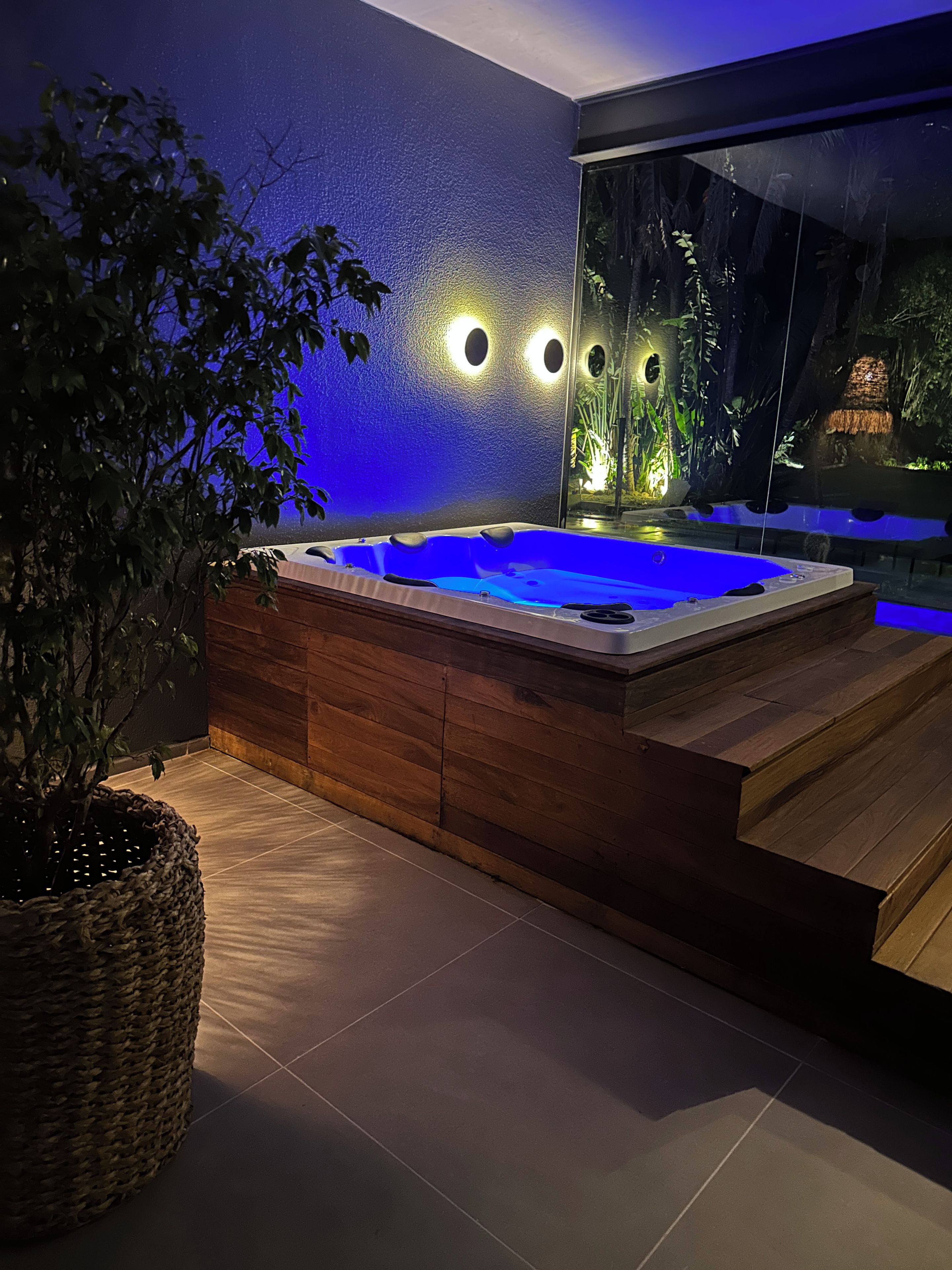 Un moderno jacuzzi interior con detalles de madera, rodeado por una pared oscura con luces circulares y un amplio ventanal con vistas a un jardín.