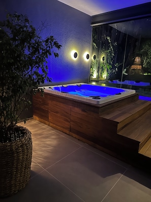 Ein modernes Indoor-Whirlpool mit Holzelementen, umgeben von einer dunklen Wand mit runden Lichtern und einem großen Fenster mit Blick auf einen Garten.