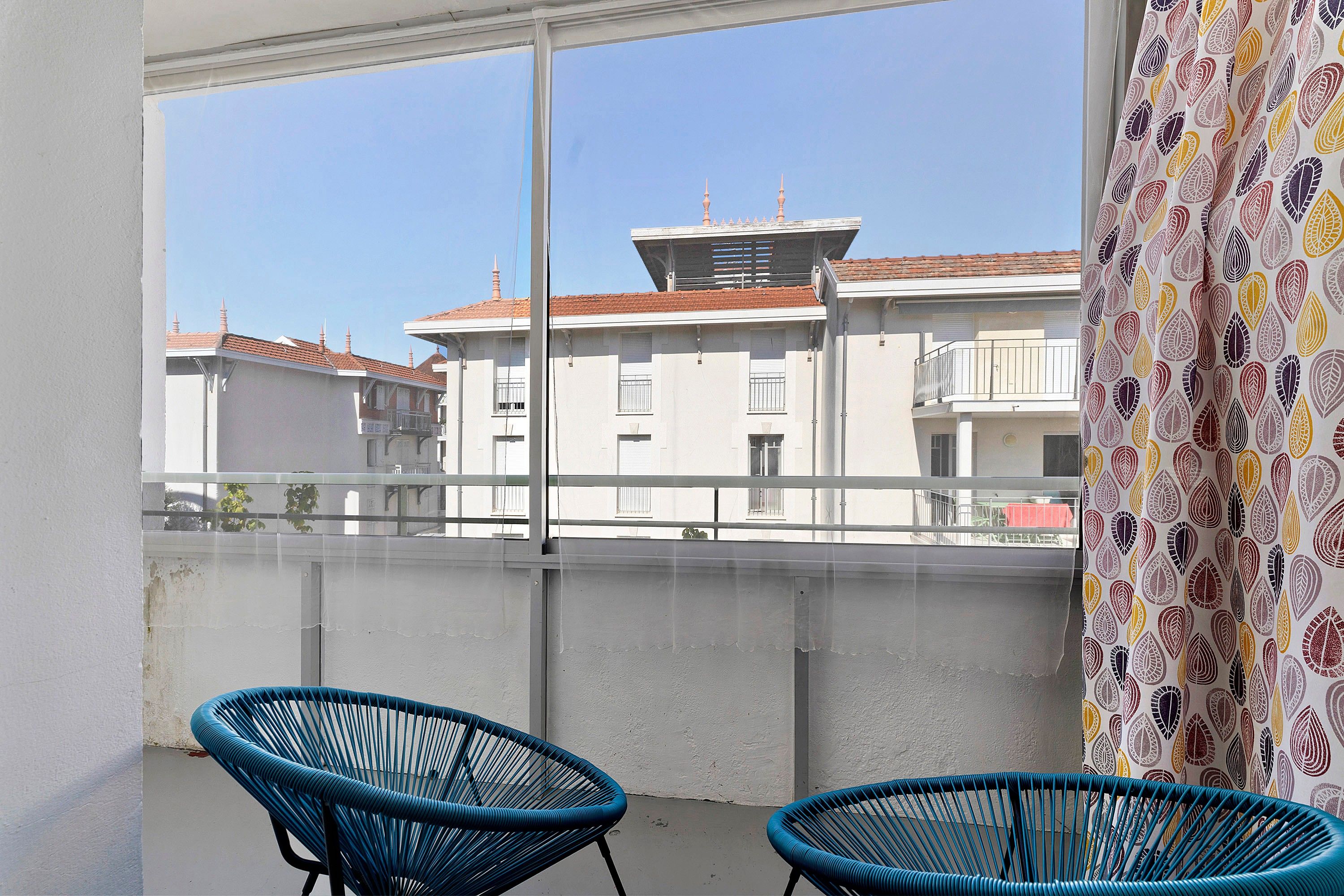 Un balcon avec deux chaises bleues, une vue sur des immeubles aux toits rouges et un rideau à motifs.