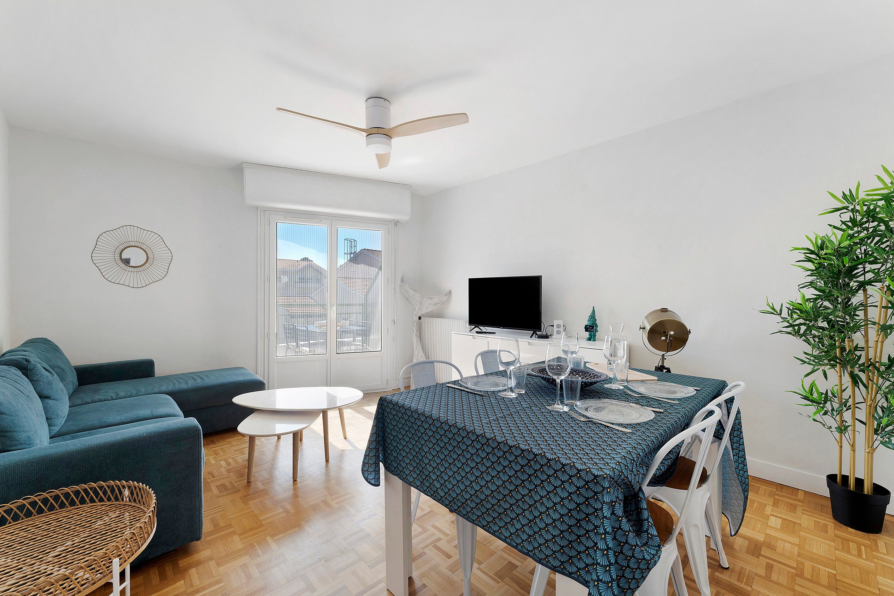 A modern living room with a blue sofa, a dining table set for four, a television, and a large window with a view of buildings.