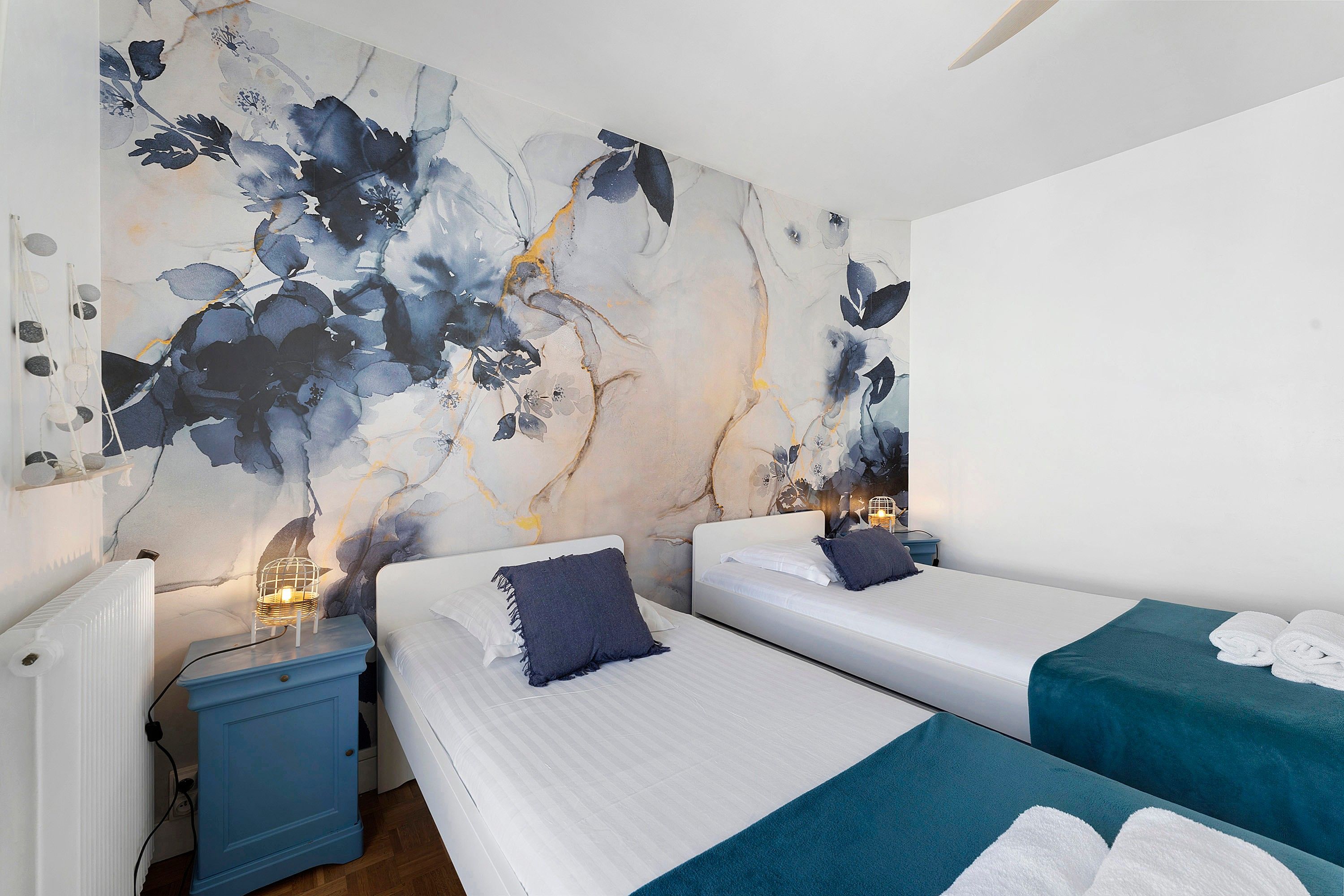 A hotel room with two beds, a nightstand, a wall with a floral mural, and a ceiling fan.