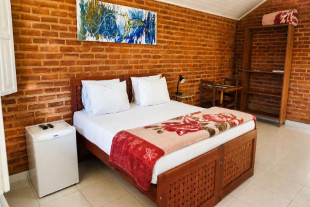 Un dormitorio con pared de ladrillo, una cama con ropa blanca, una manta roja, un electrodoméstico pequeño blanco y un armario de madera.