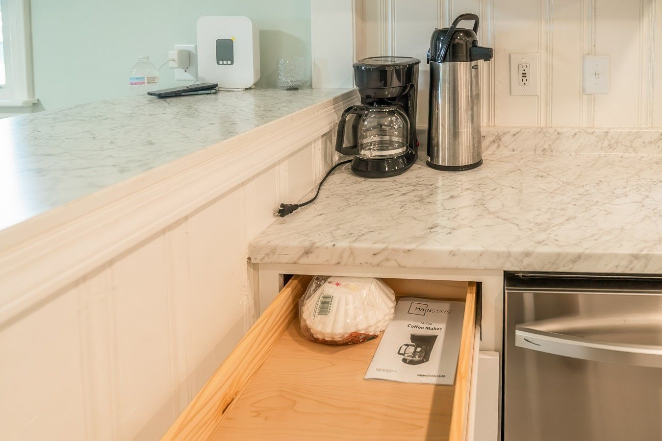 A kitchen with a marble countertop, a coffee maker, a kettle, and a drawer with a coffee maker box.