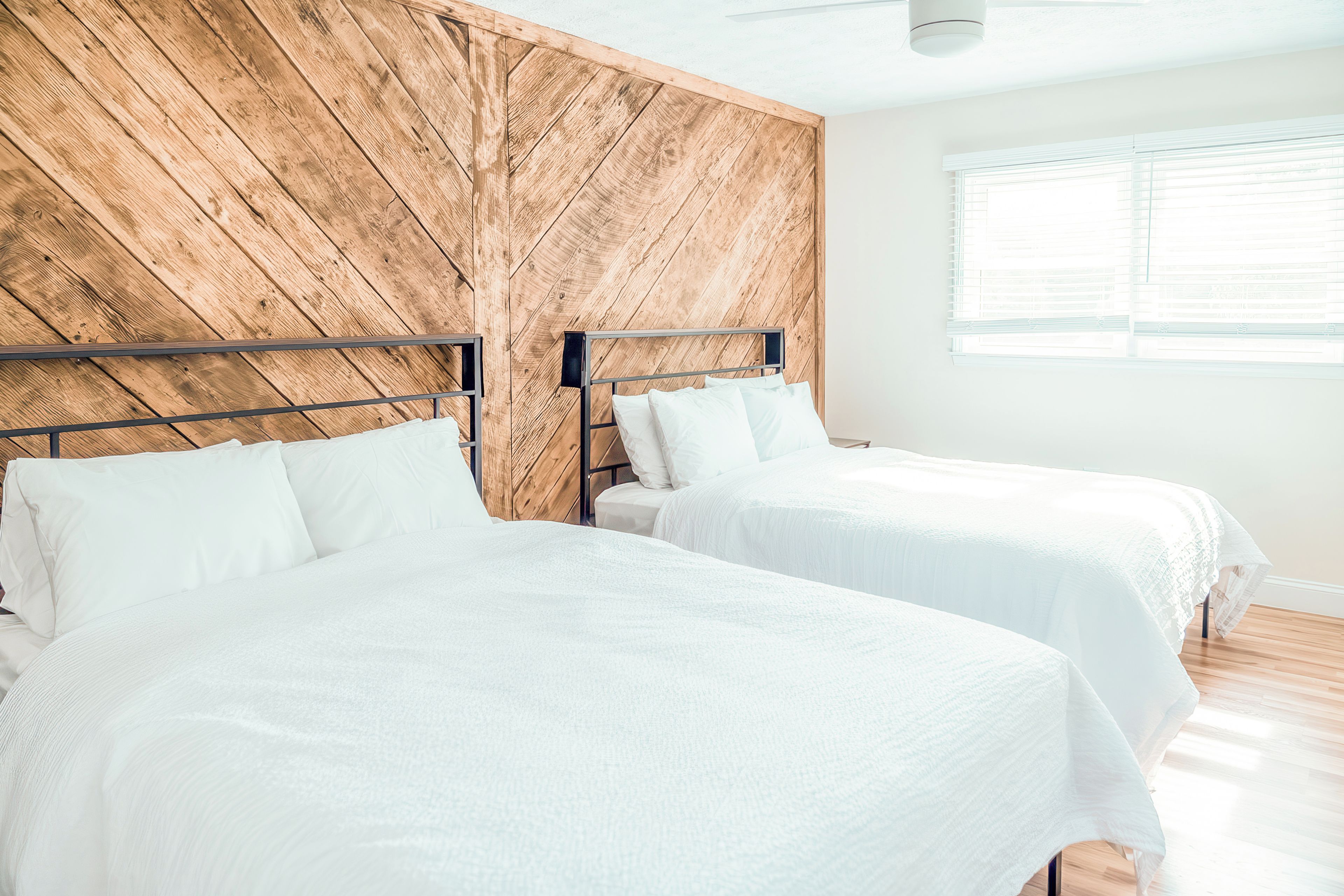 A bedroom with two beds, wooden headboard, white bedding, and a window with blinds.