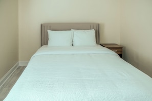 Un llit ben fet amb capçalera, una taula de nit de fusta i una paret sense decorar.