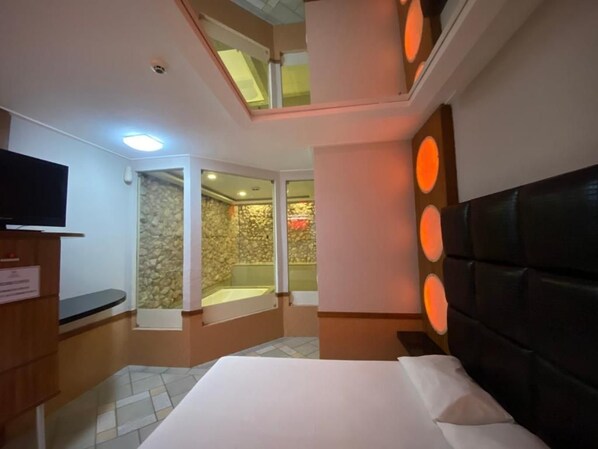 A hotel room with a bed, a television, a stone wall, a bathtub, and a skylight. - Drops Motel Palhoça (Palhoça)