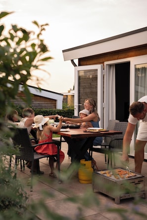 Una familia cenando al aire libre en una mesa con una parrilla, rodeada de sillas y un edificio moderno al fondo.