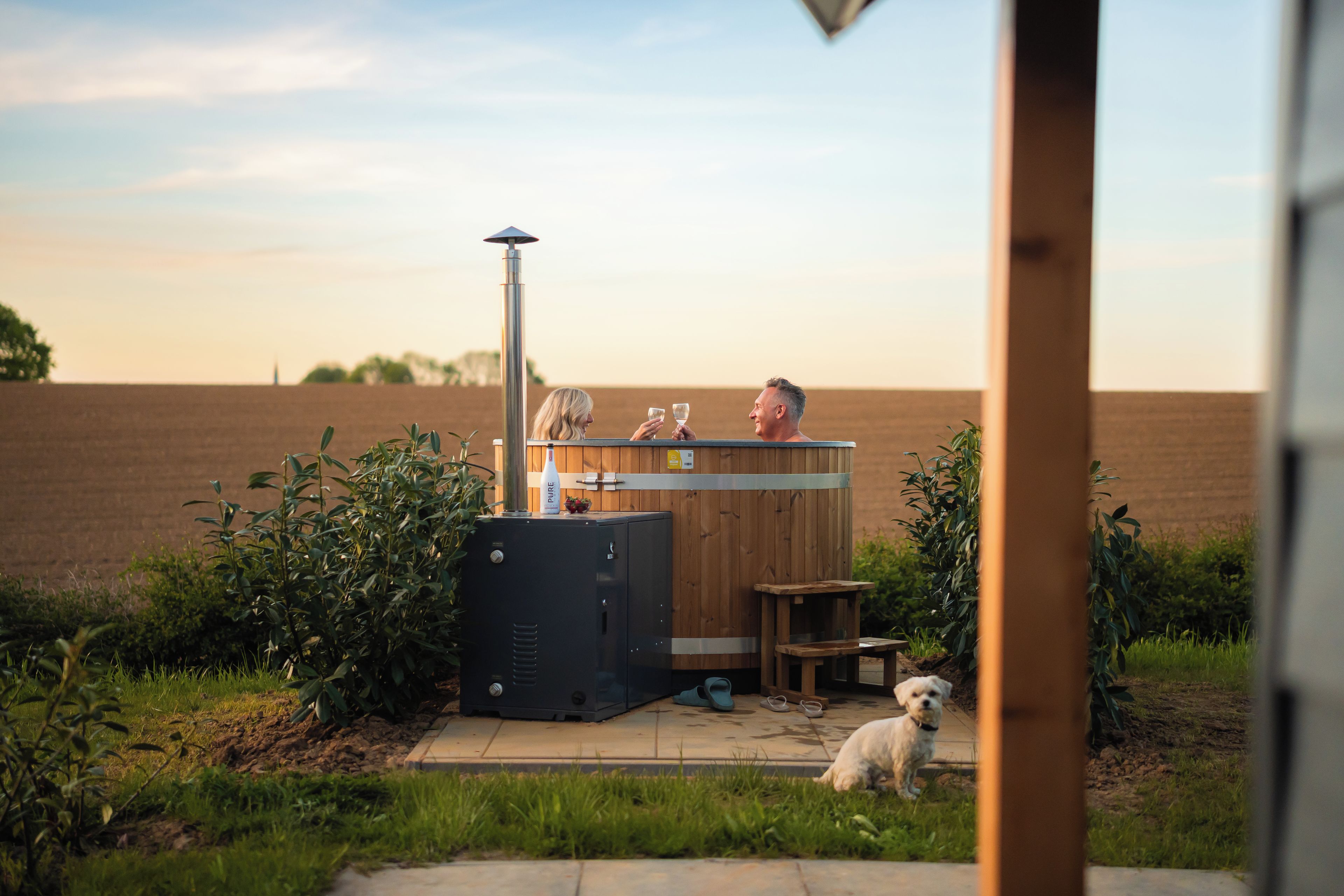 Un bain à remous en bois avec un réchauffeur en métal, une petite table en bois et un chien dans un espace herbeux.