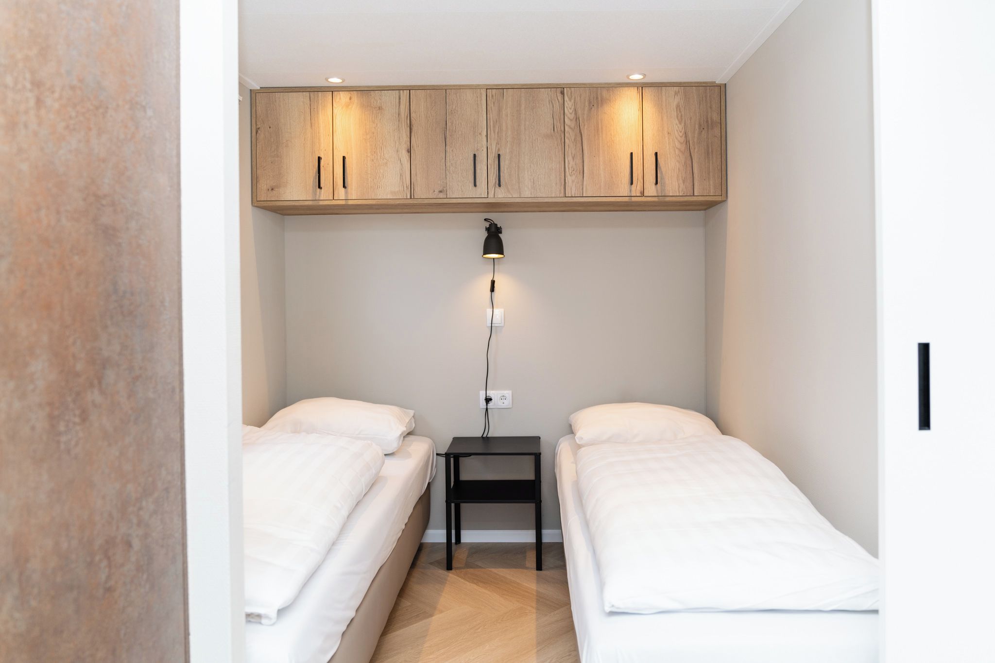 Een kleine, moderne slaapkamer met twee eenpersoonsbedden, een houten garderobe en een nachtkastje met een lamp.
