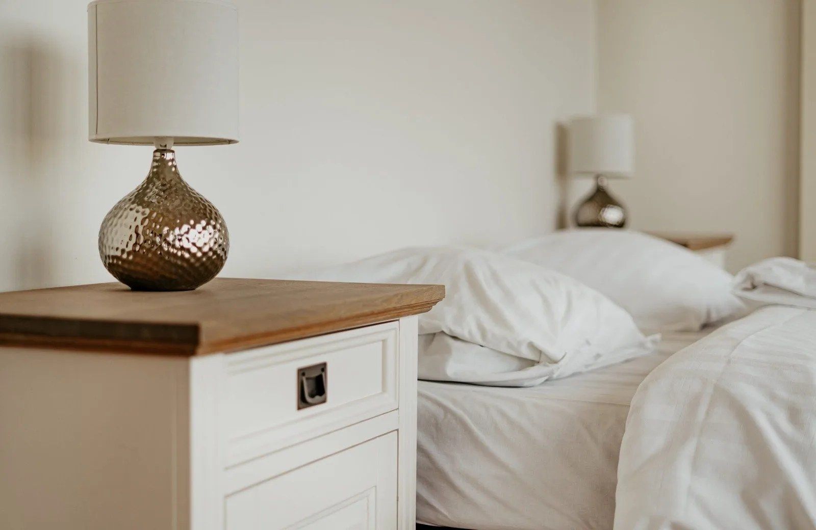 Una cama bien hecha con sábanas blancas y una mesita de noche de madera con una lámpara.