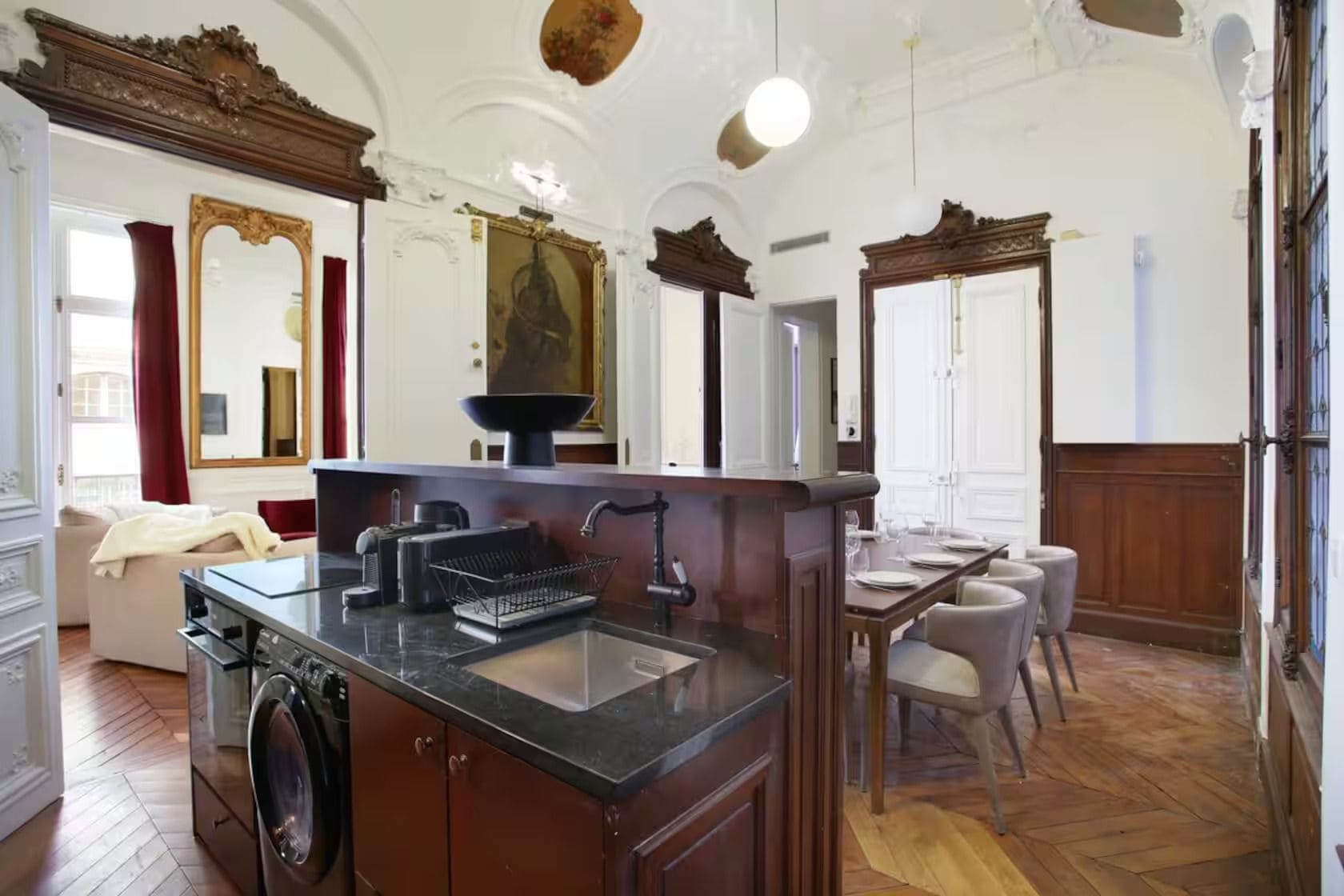 A spacious living area with a large window, a dining table set for four, and a kitchen with a washing machine.
