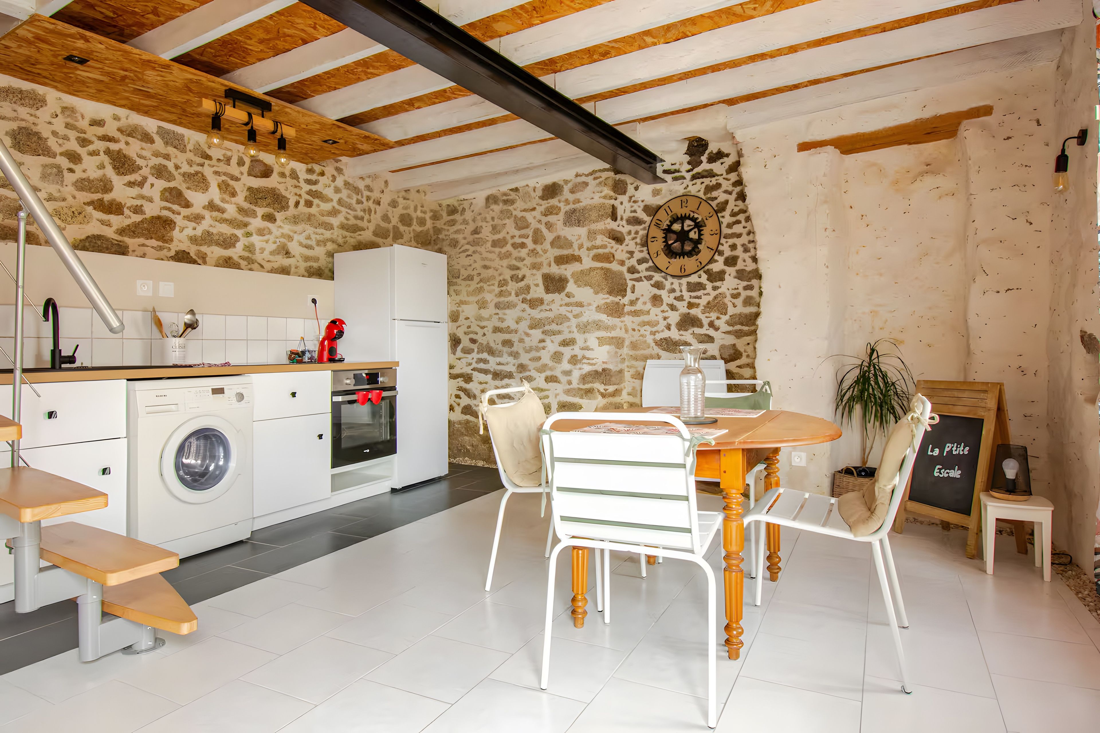 Une cuisine avec un mur en pierre, des poutres en bois au plafond, un lave-linge et un coin repas avec une table et des chaises.