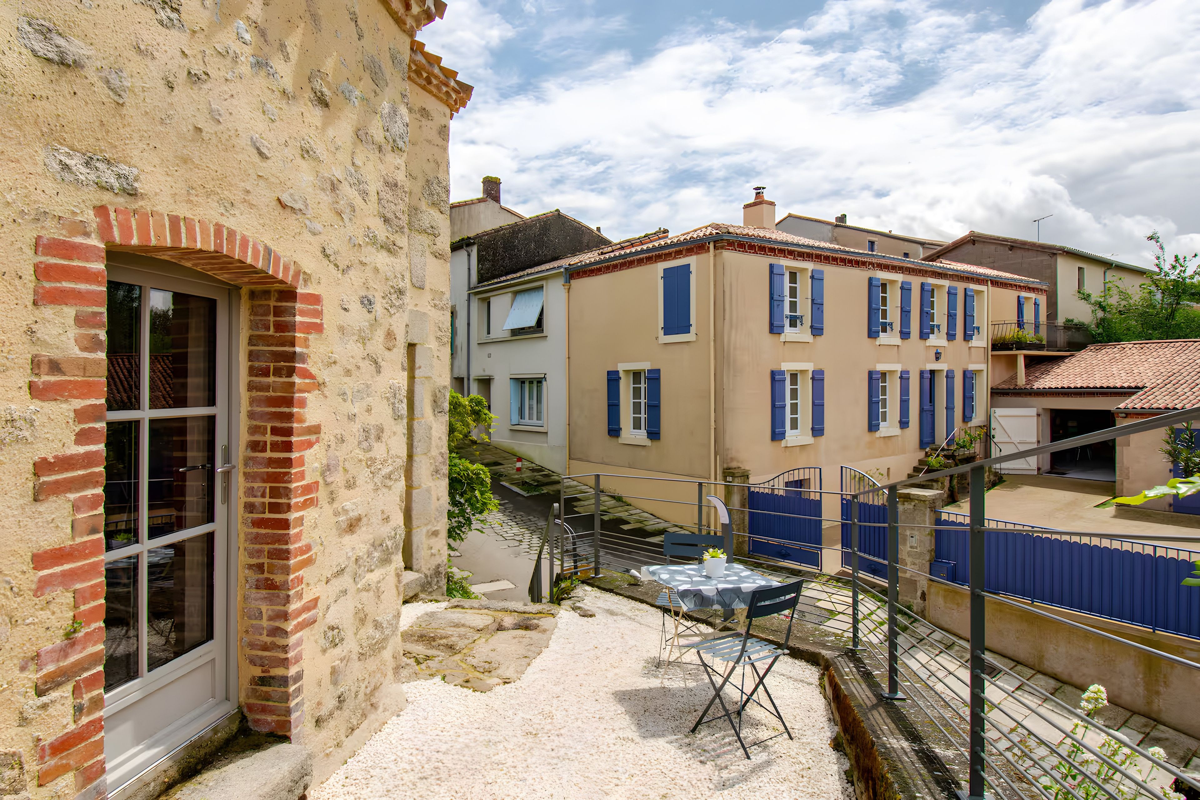 A courtyard with a table and chairs, a brick wall, and a building with blue shutters.
