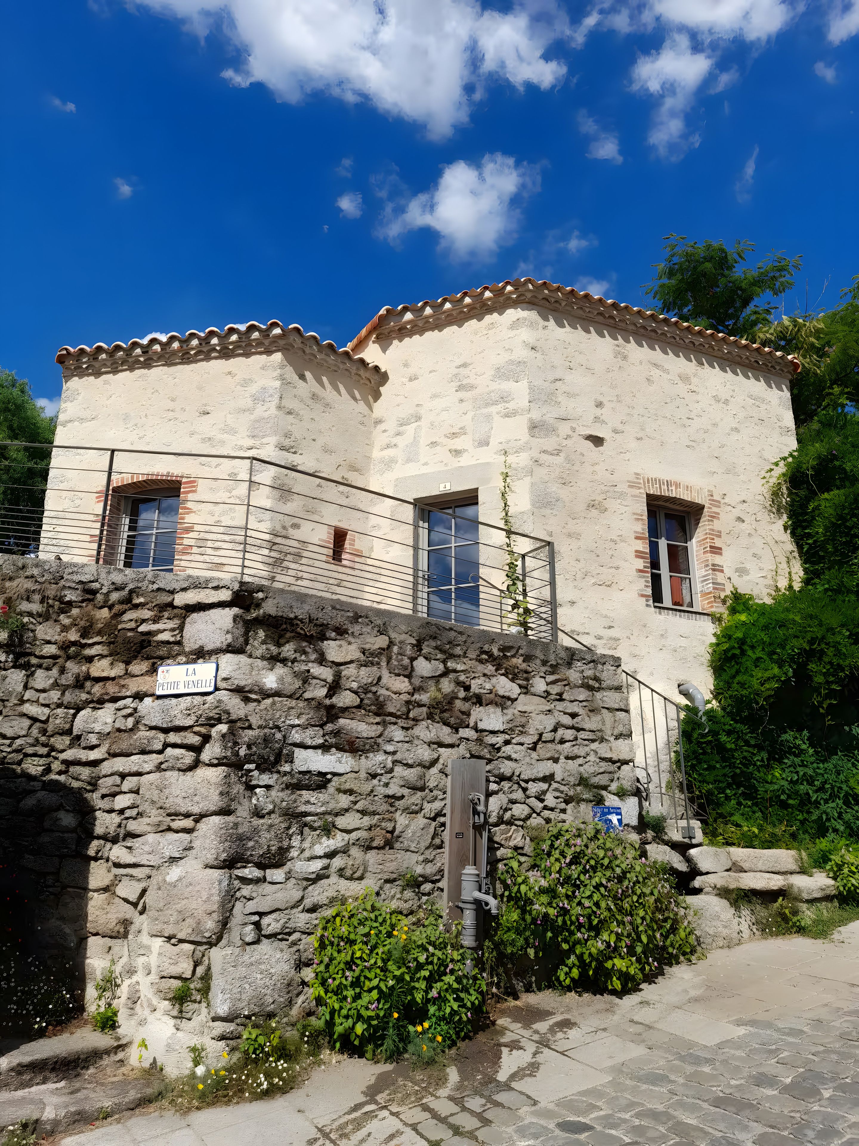 Un bâtiment en pierre avec un toit en tuiles, une rambarde en métal et un mur en pierre muni d’un robinet.