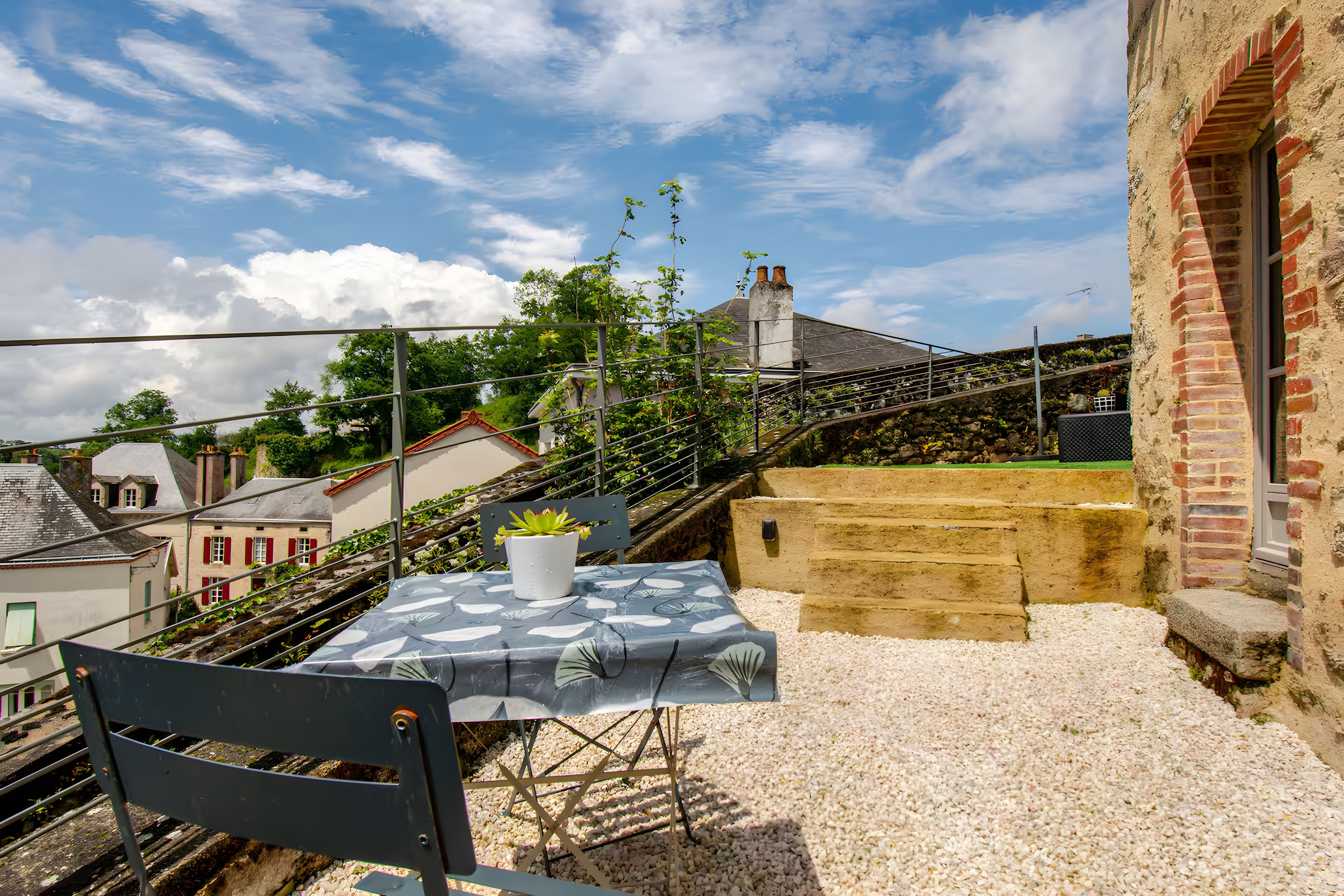 A terrace with a table and chair, a potted plant, and a view of buildings and trees.