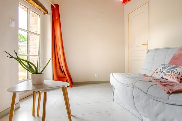 Une pièce avec un canapé blanc, une petite table sur laquelle est posée une plante, une fenêtre avec des rideaux et une porte.