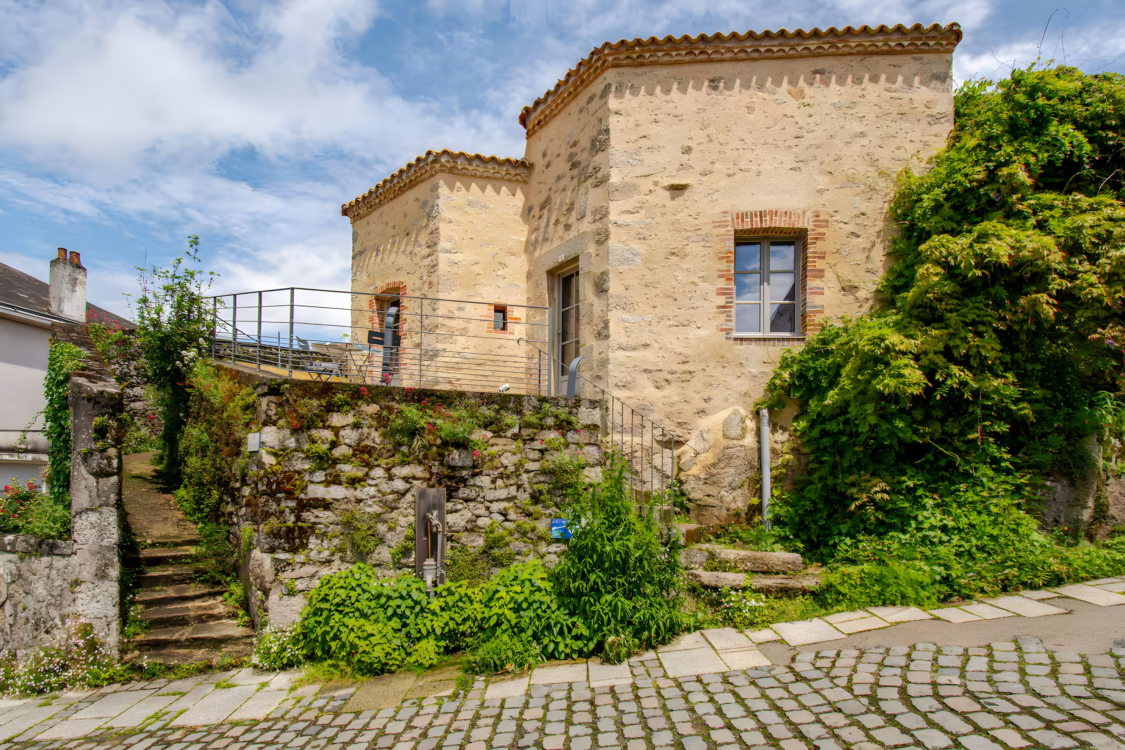 A stone building with a traditional design, a staircase leading to a door, and a garden area with greenery.