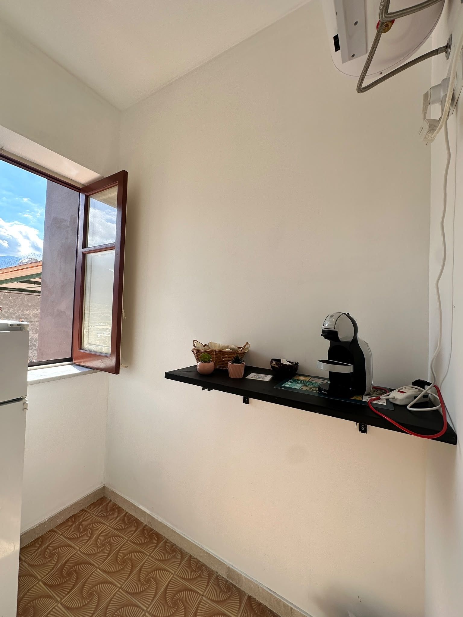 A small kitchen with a window, a shelf with kitchen appliances, and a patterned floor.