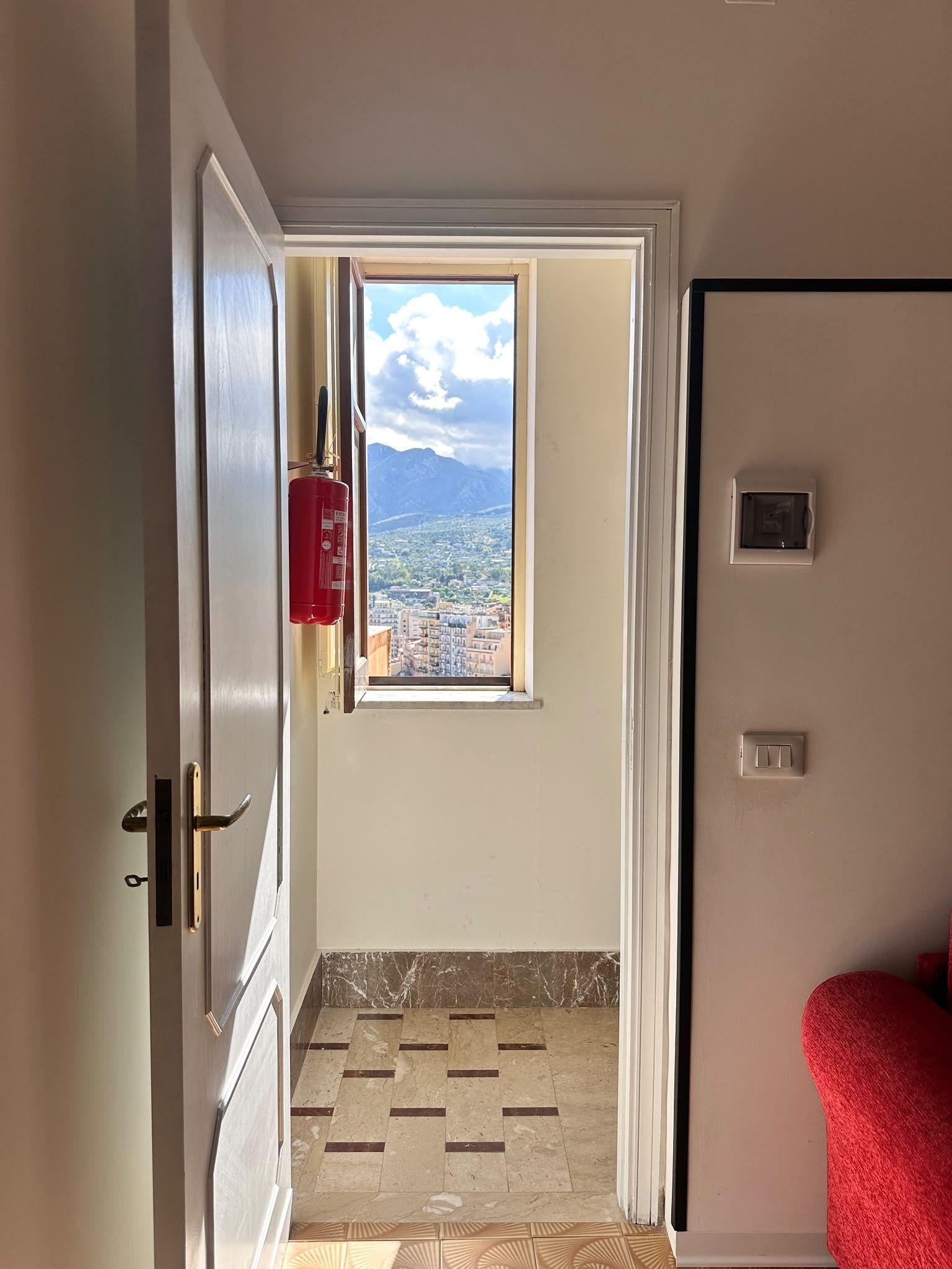 A room with a door leading to a balcony with a city view, a red fire extinguisher, and a tiled floor.