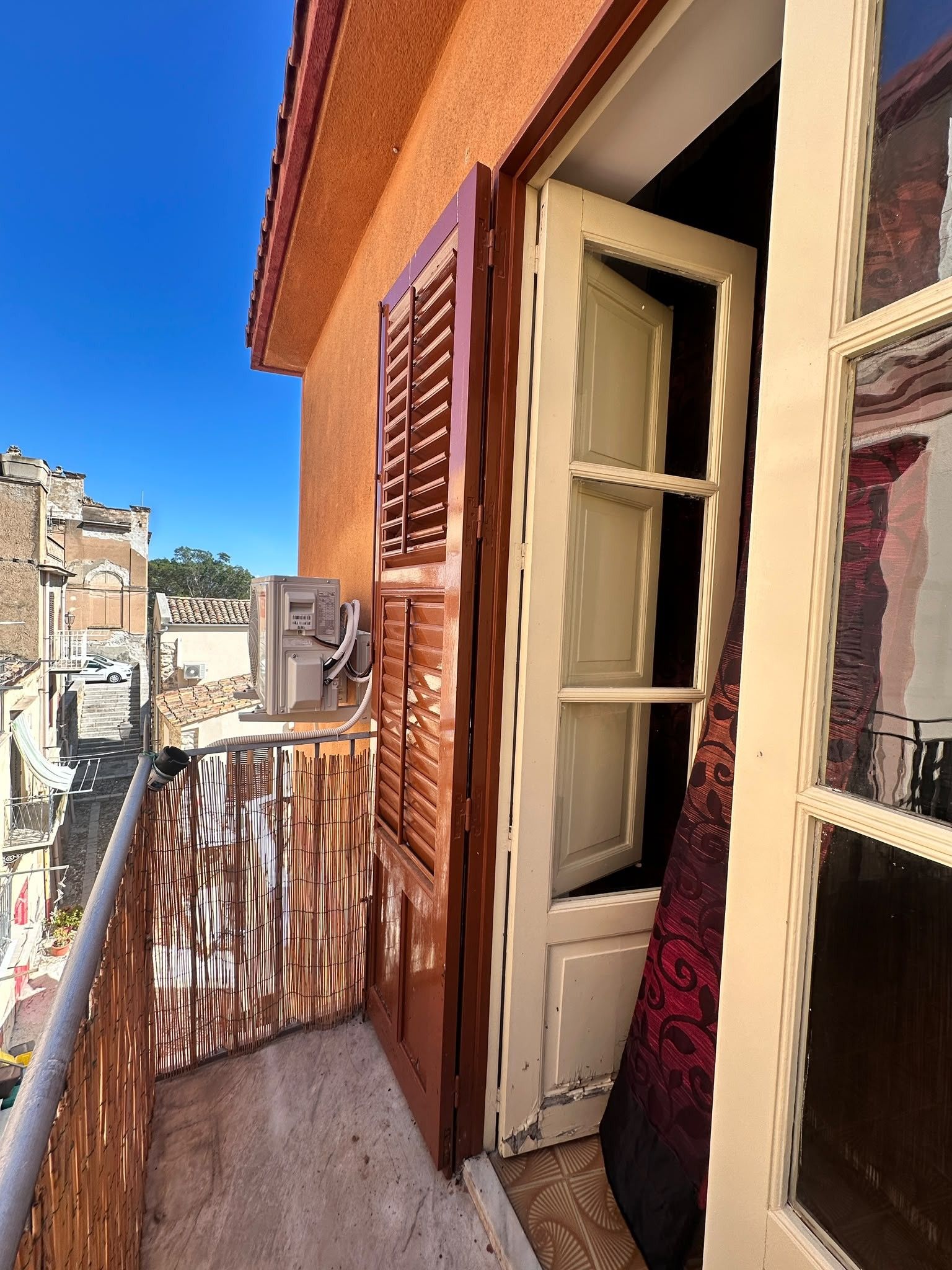 Ein Balkon mit Blick auf angrenzende Gebäude, eine Tür mit einem gemusterten Vorhang und eine Klimaanlage.