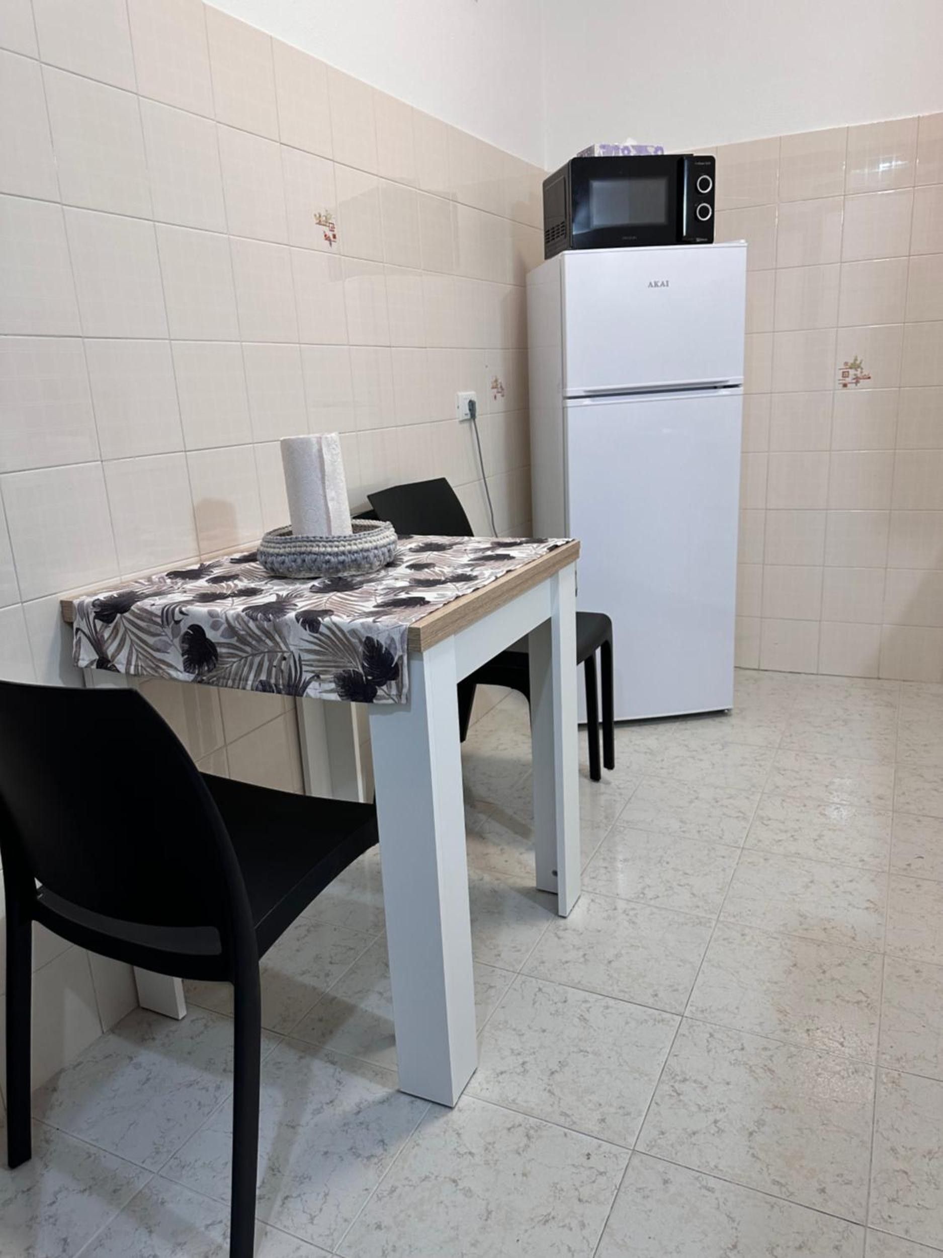 Eine kleine Küche mit einem weißen Kühlschrank, einer Mikrowelle darauf, einem Tisch mit einem gemusterten Tischläufer und zwei schwarzen Stühlen.