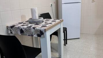 Eine kleine Küche mit einem weißen Kühlschrank, einer Mikrowelle darauf, einem Tisch mit einem gemusterten Tischläufer und zwei schwarzen Stühlen.