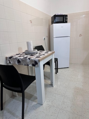 Eine kleine Küche mit einem weißen Kühlschrank, einer Mikrowelle darauf, einem Tisch mit einem gemusterten Tischläufer und zwei schwarzen Stühlen.