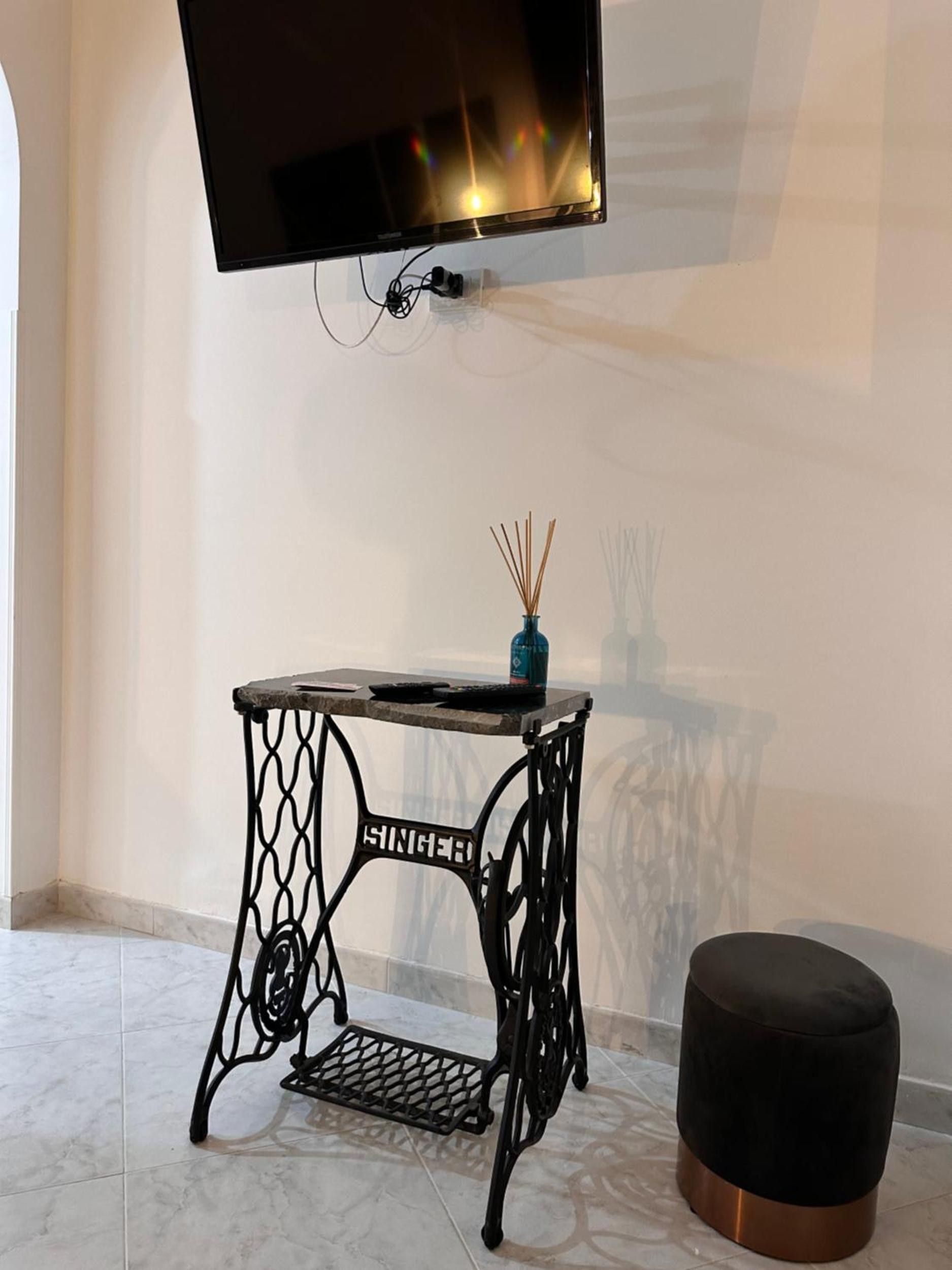 Ein Zimmer mit an der Wand montiertem Fernseher, einem kleinen Tisch mit einer Vase und einem schwarzen Ottomanen.