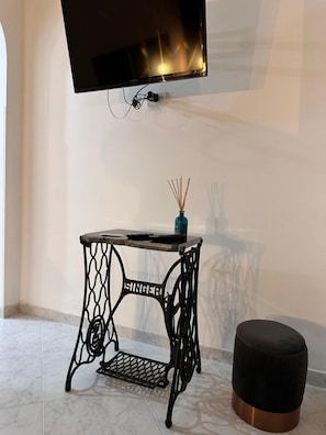 Ein Zimmer mit an der Wand montiertem Fernseher, einem kleinen Tisch mit einer Vase und einem schwarzen Ottomanen.
