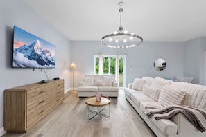 A modern living room with a large flat-screen TV, a wooden cabinet, a round coffee table, a white sofa, and a chandelier.