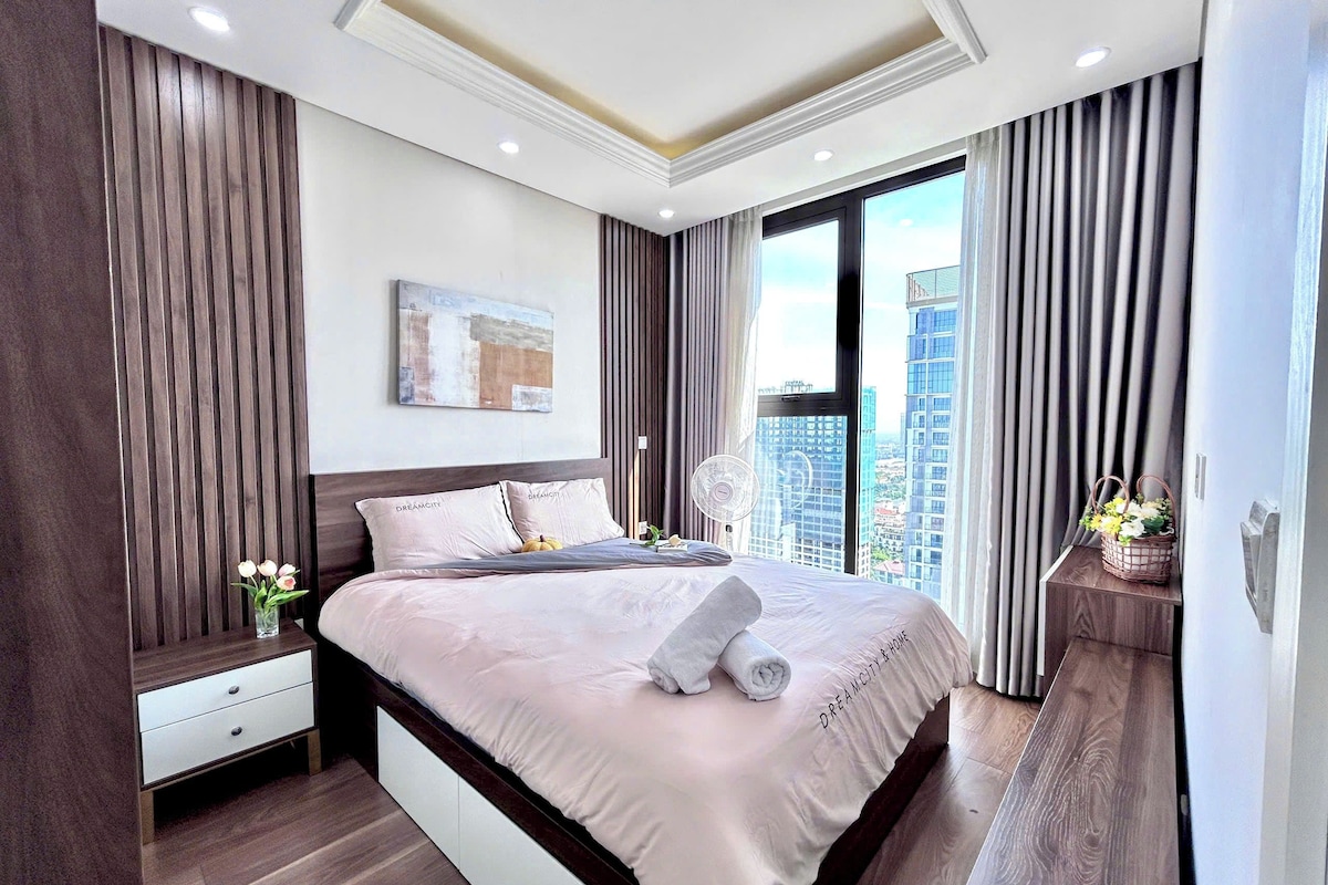 A modern hotel room with a large bed, a bedside table, a wall-mounted artwork, and a view of the cityscape through a large window.