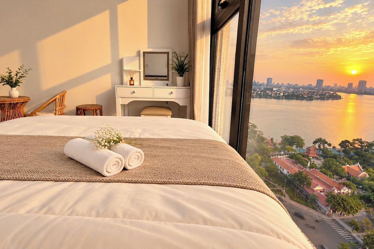 A hotel room with a bed, a desk, and a view of a cityscape at sunset.
