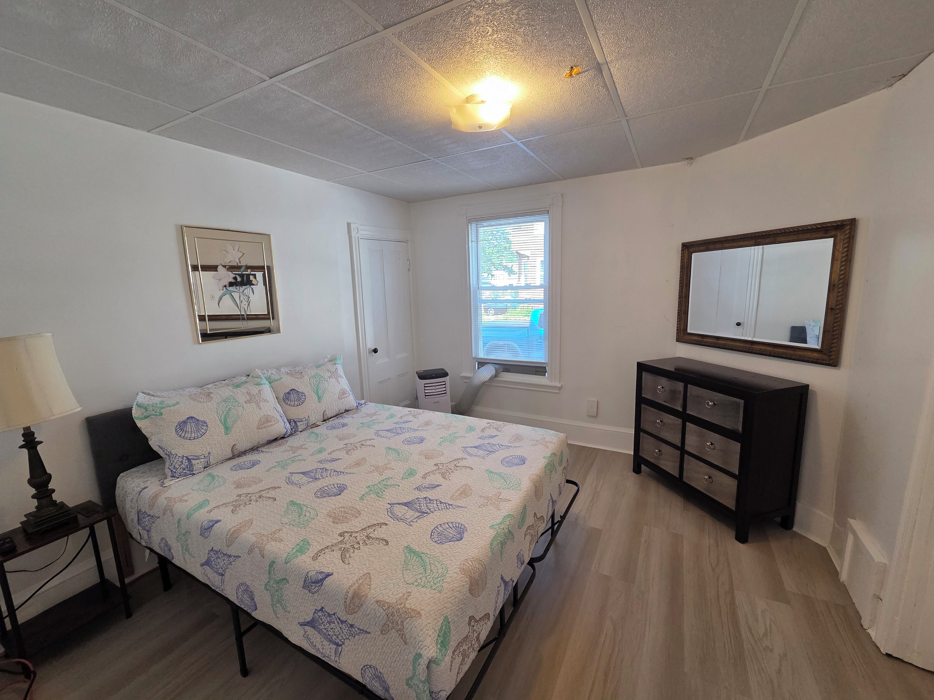 Une chambre à coucher comprenant un lit, une commode, une table de chevet, une lampe, une fenêtre avec des stores et un luminaire au plafond.