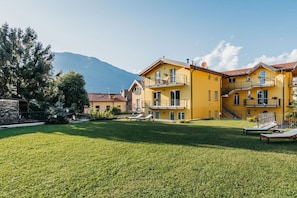 A yellow building with balconies, a grassy courtyard, and a mountain in the background. - New Listing (Dervio)