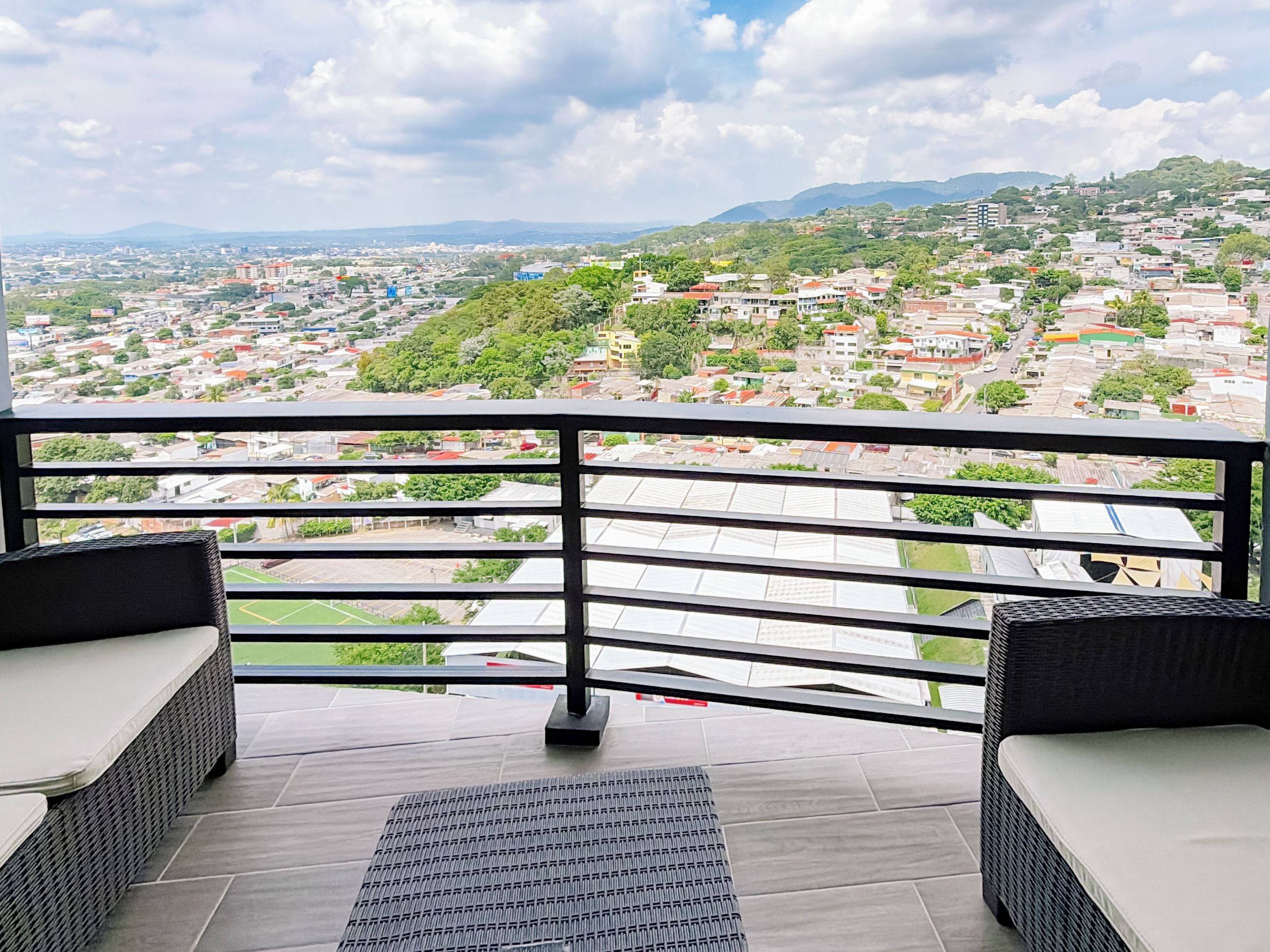 A balcony with a view of a cityscape, including buildings and greenery.
