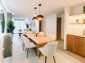 A modern dining area with a wooden table, white chairs, pendant lighting, and a kitchen area with a wooden countertop. - Urban Nest Lomas, 3 hab., vistas panorámicas, AC, balcón y 2 parqueos (San Salvador)