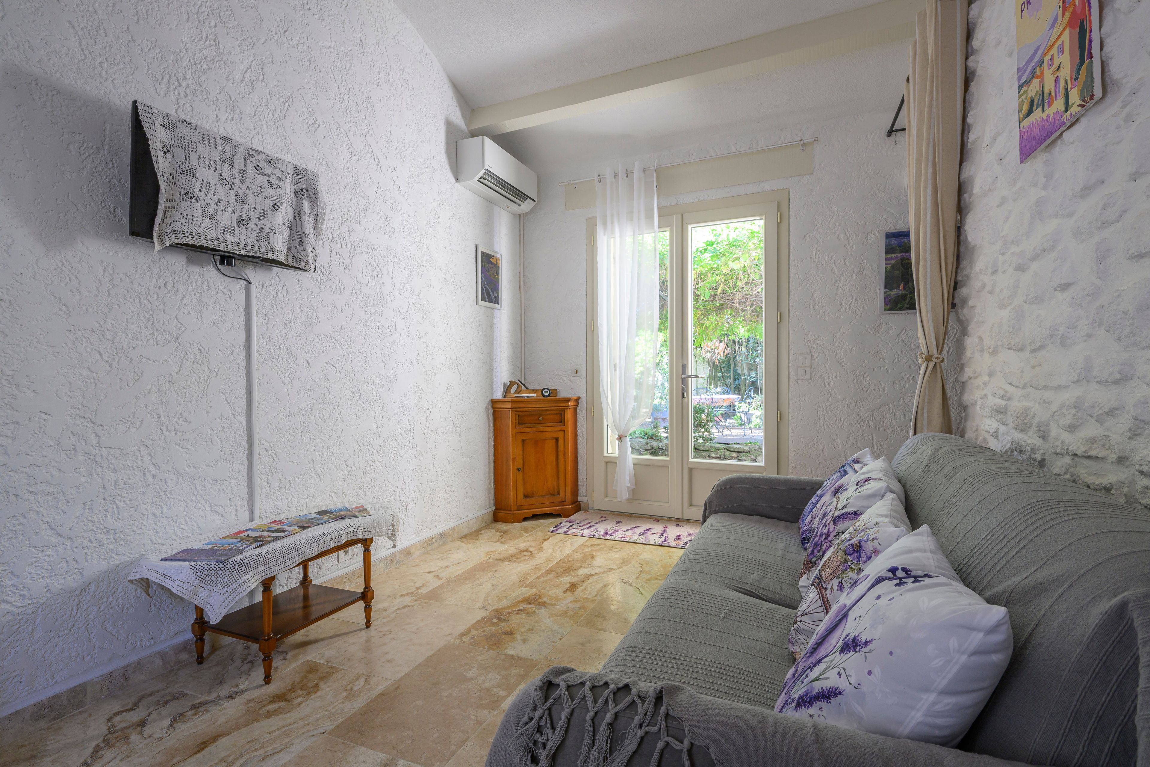Un salon avec un canapé, une petite table en bois, un téléviseur fixé au mur et une fenêtre avec des rideaux.