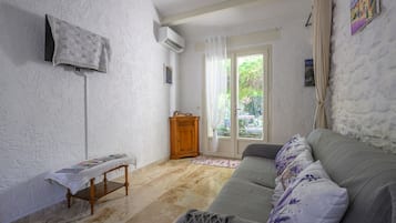 Un salon avec un canapé, une petite table en bois, un téléviseur fixé au mur et une fenêtre avec des rideaux.