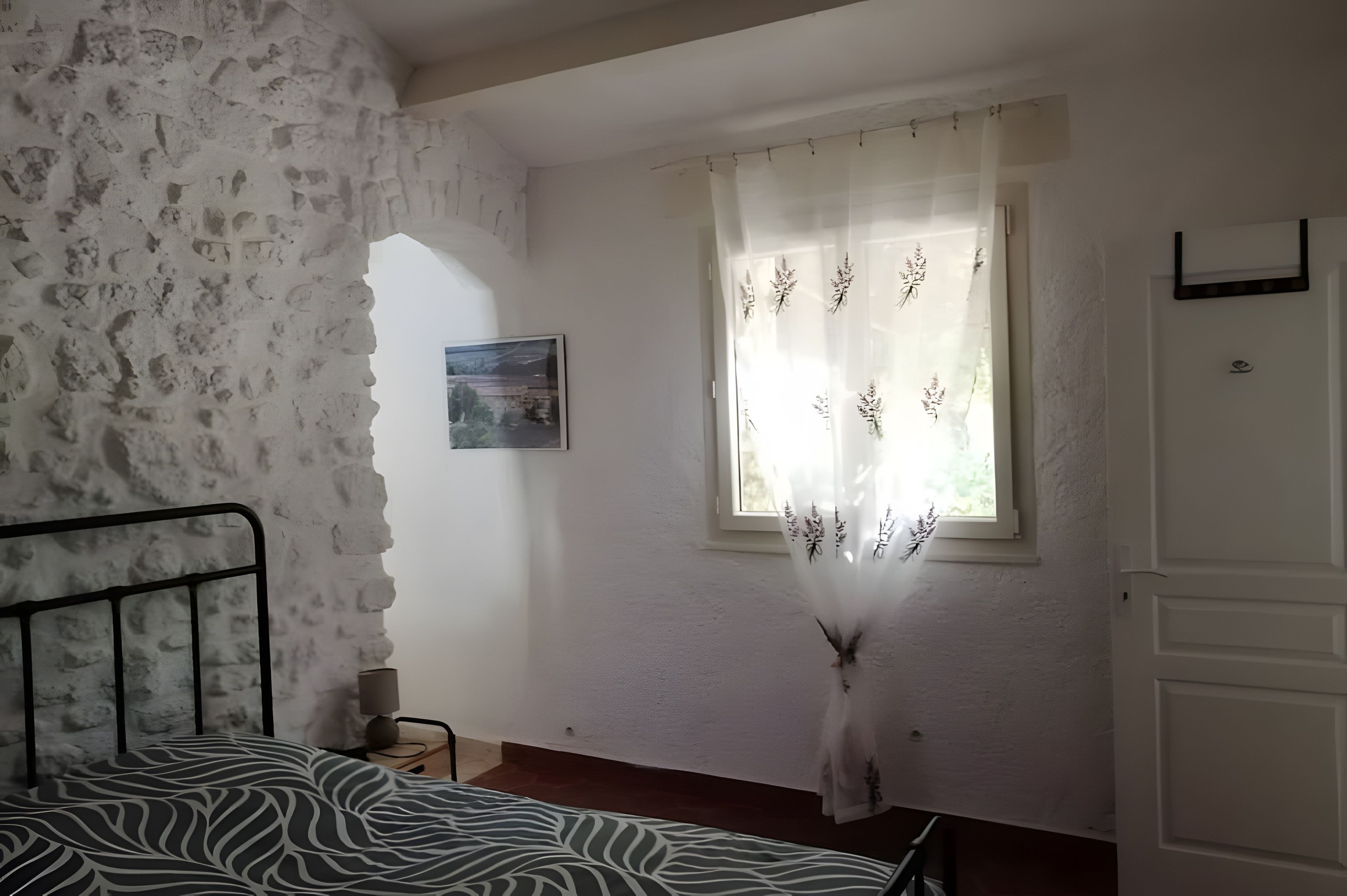 Une chambre avec un mur en pierre, un lit recouvert d’une couvre-lit à motifs zébrés, une fenêtre ornée de rideaux à fleurs et une petite table de chevet avec une lampe.