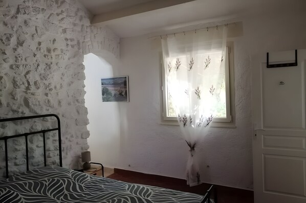 A bedroom with a stone wall, a bed with a zebra-patterned bedspread, a window with floral curtains, and a small bedside table with a lamp. - A fully renovated former sheep barn, located in the petite luberon region (Cheval-Blanc)