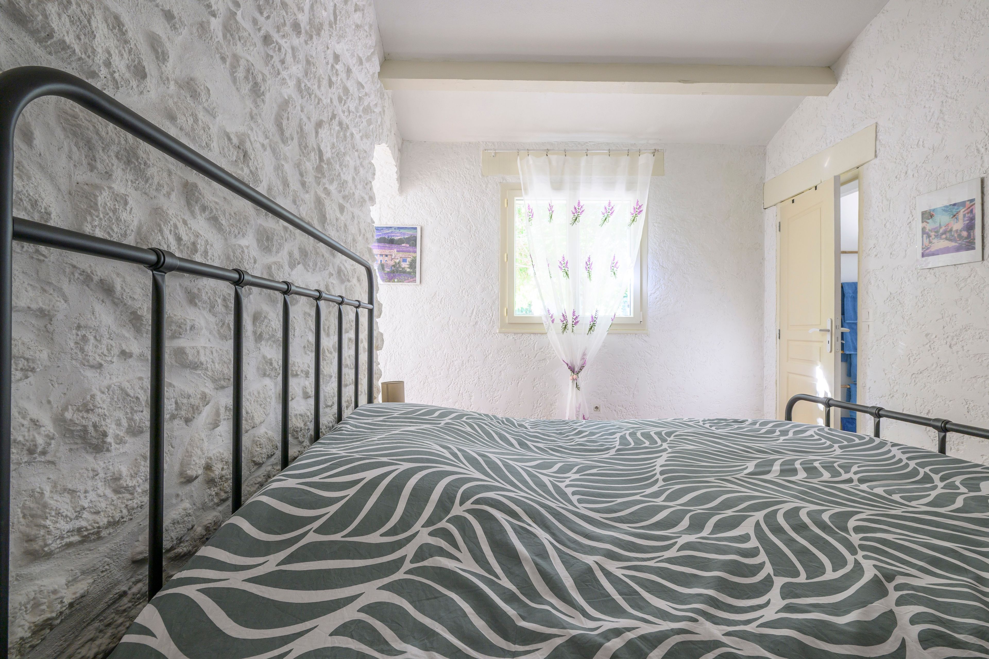 Une chambre à coucher avec un lit, une fenêtre ornée de rideaux à motifs floraux et un mur blanc à la texture particulière.