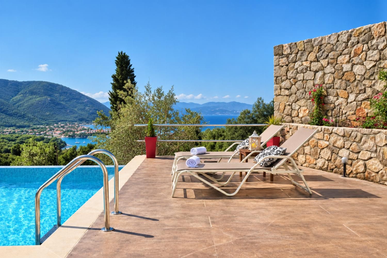 A poolside area with a stone wall, lounge chairs, and a view of the sea and mountains.