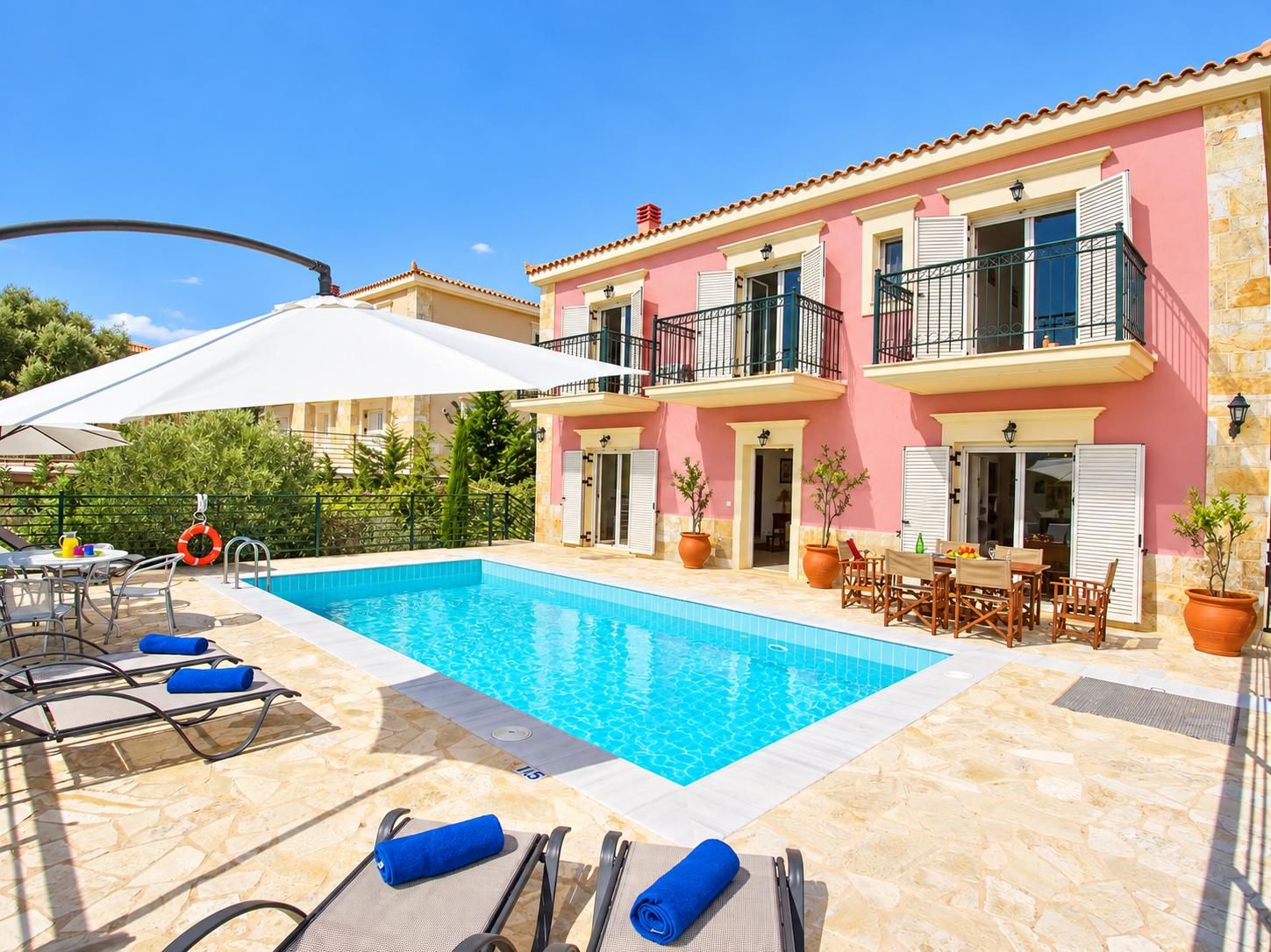 A swimming pool with lounge chairs and umbrellas, a patio area with outdoor furniture, and a pink building with balconies.