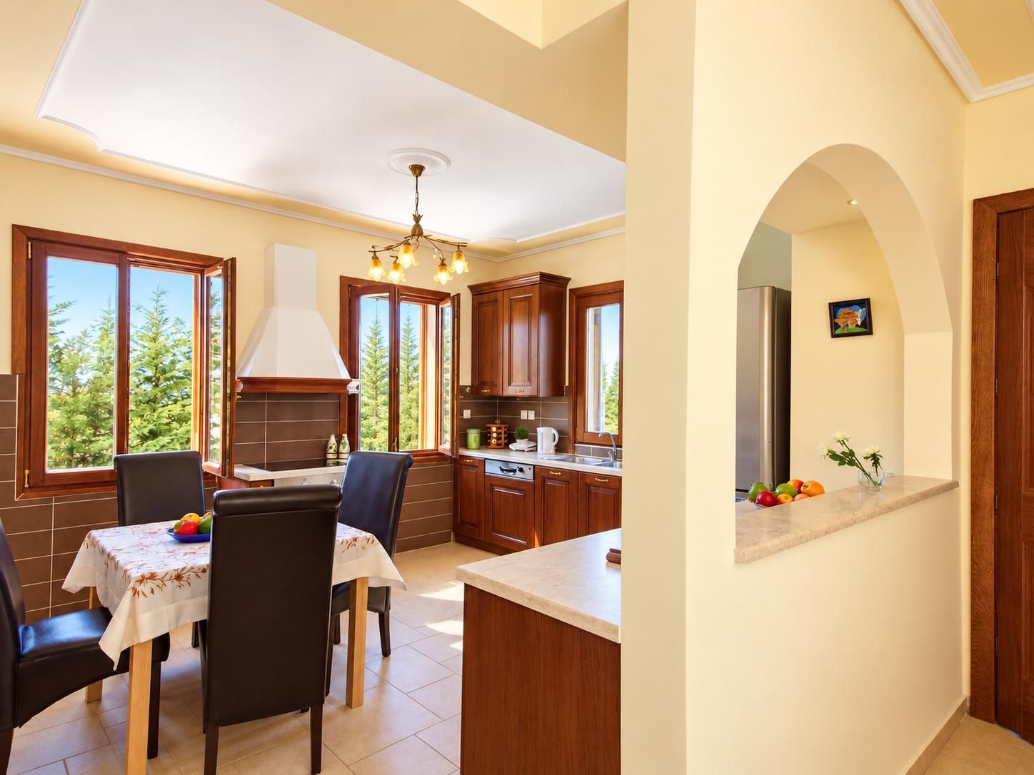 A modern kitchen with wooden cabinets, a dining area with a table and chairs, and a view of trees through large windows.