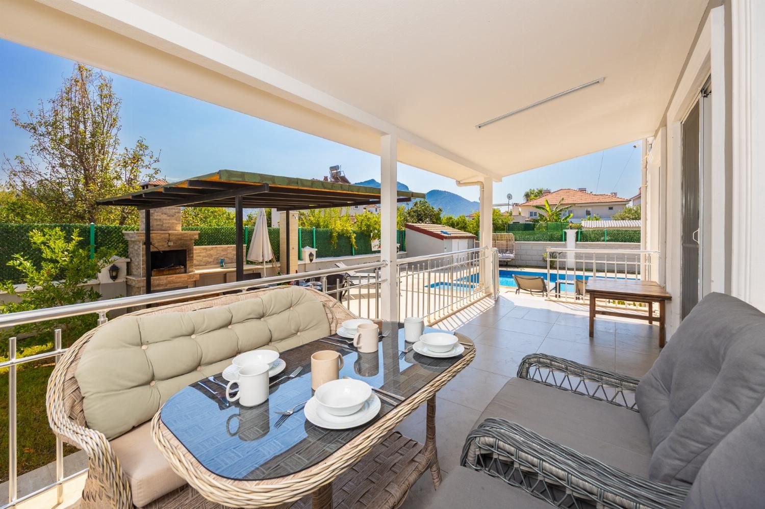 Ein überdachter Essbereich im Freien mit Tisch für zwei Personen, einer gepolsterten Couch und Blick auf einen Swimmingpool und Berge im Hintergrund.