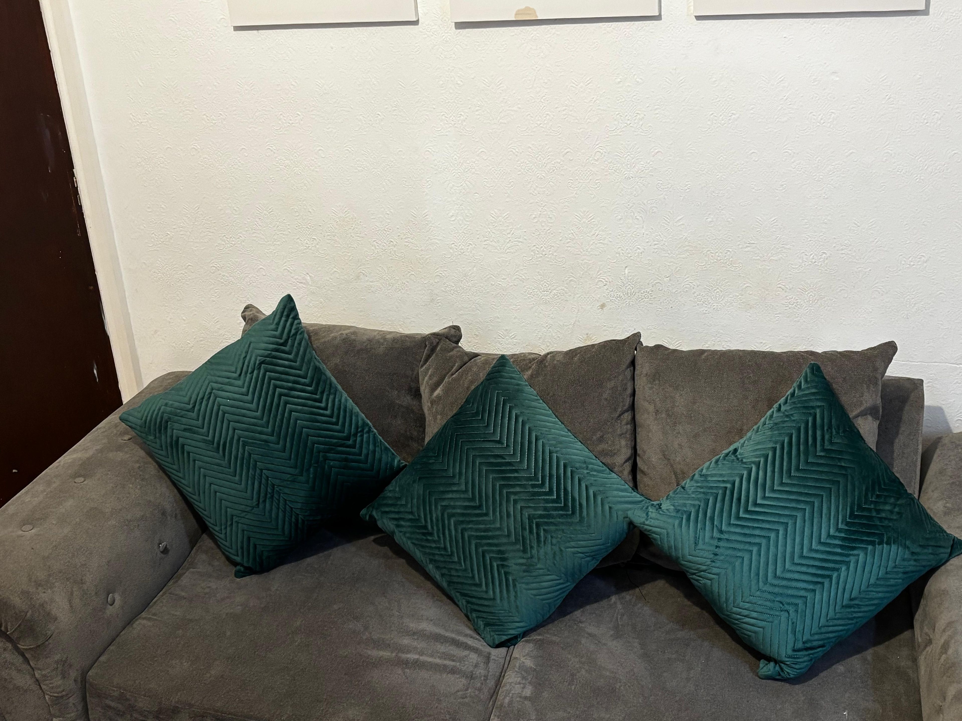 A grey sofa with three teal pillows against a white wall with a subtle pattern.