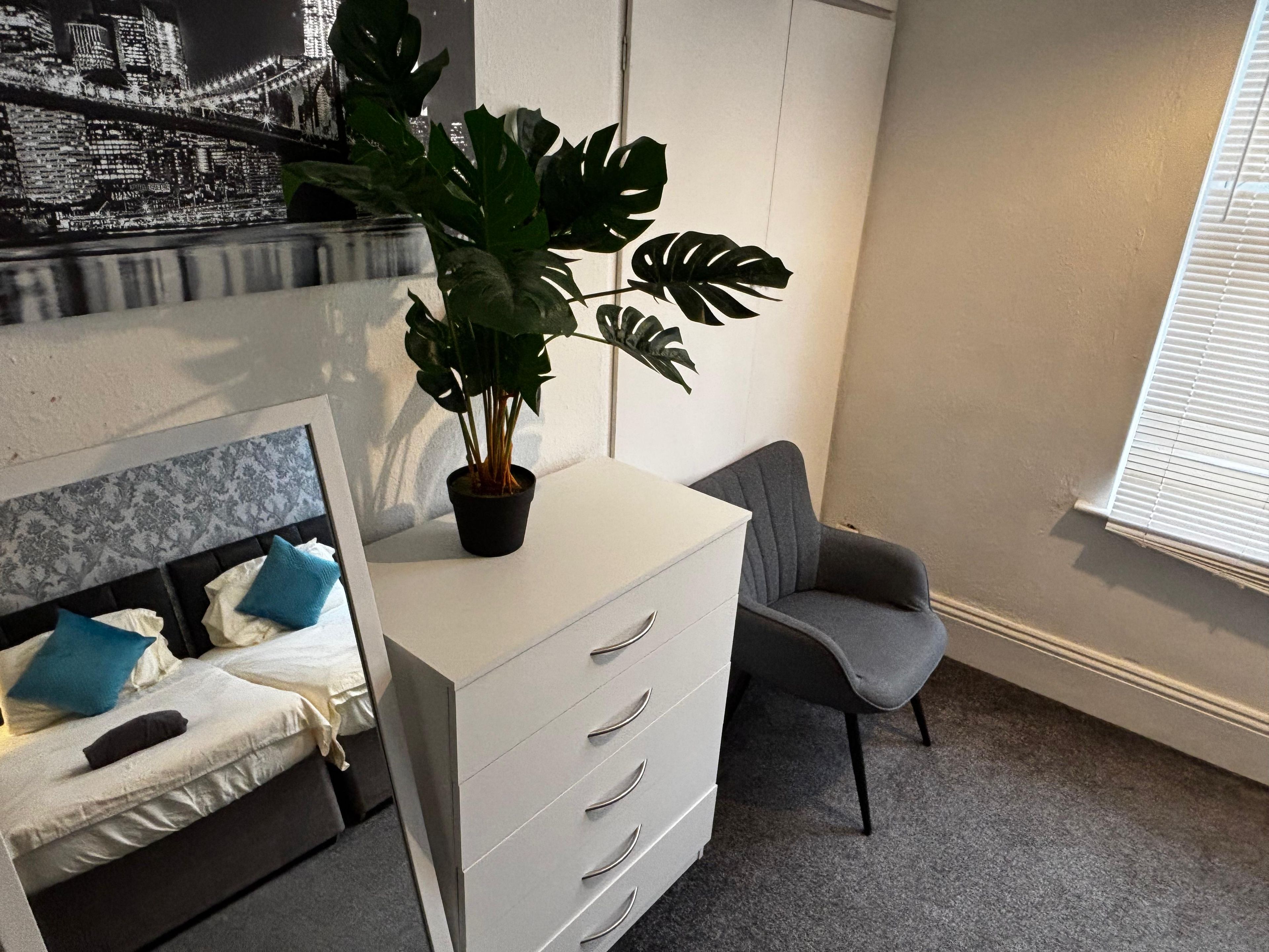 A bedroom with a bed, a dresser, a chair, and a potted plant.