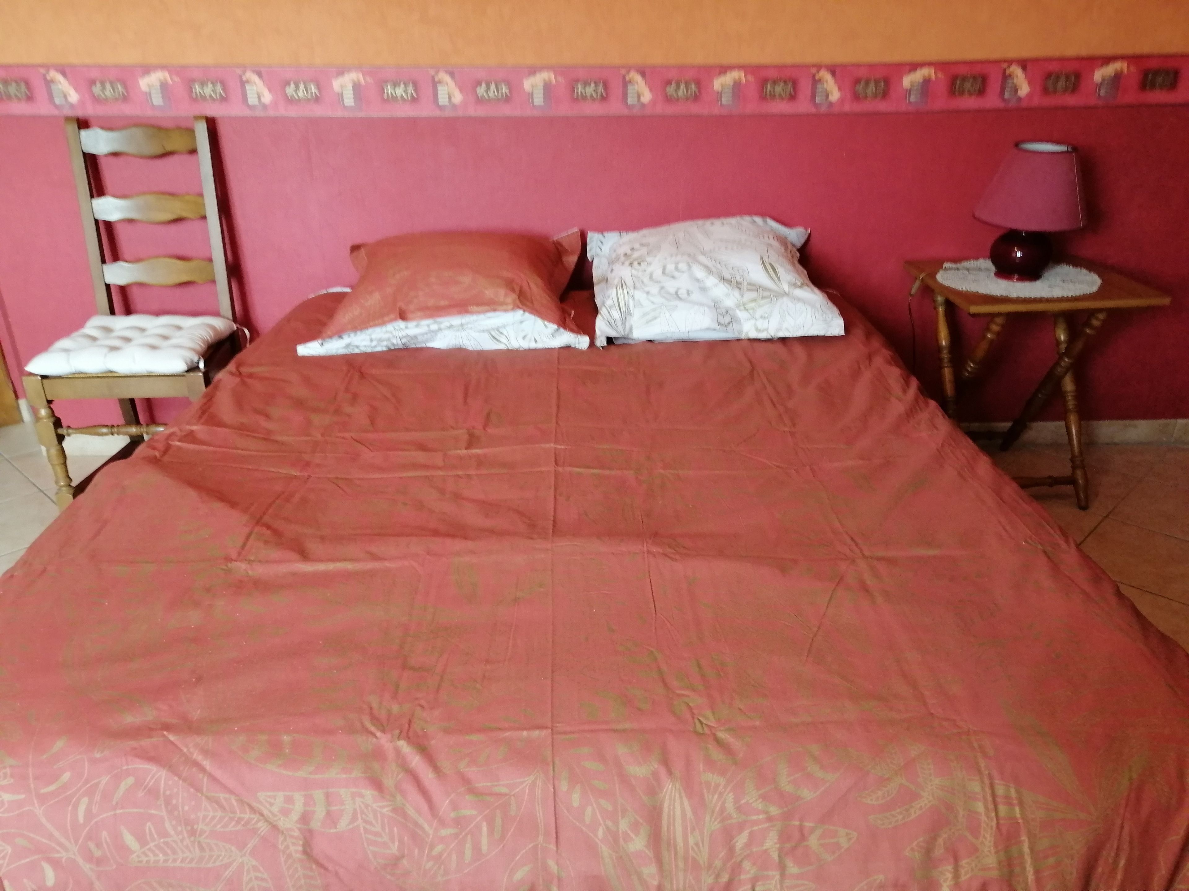 Een bed met een rode sprei, een houten stoel, een nachtkastje met een lamp, en een behang met een patroon.