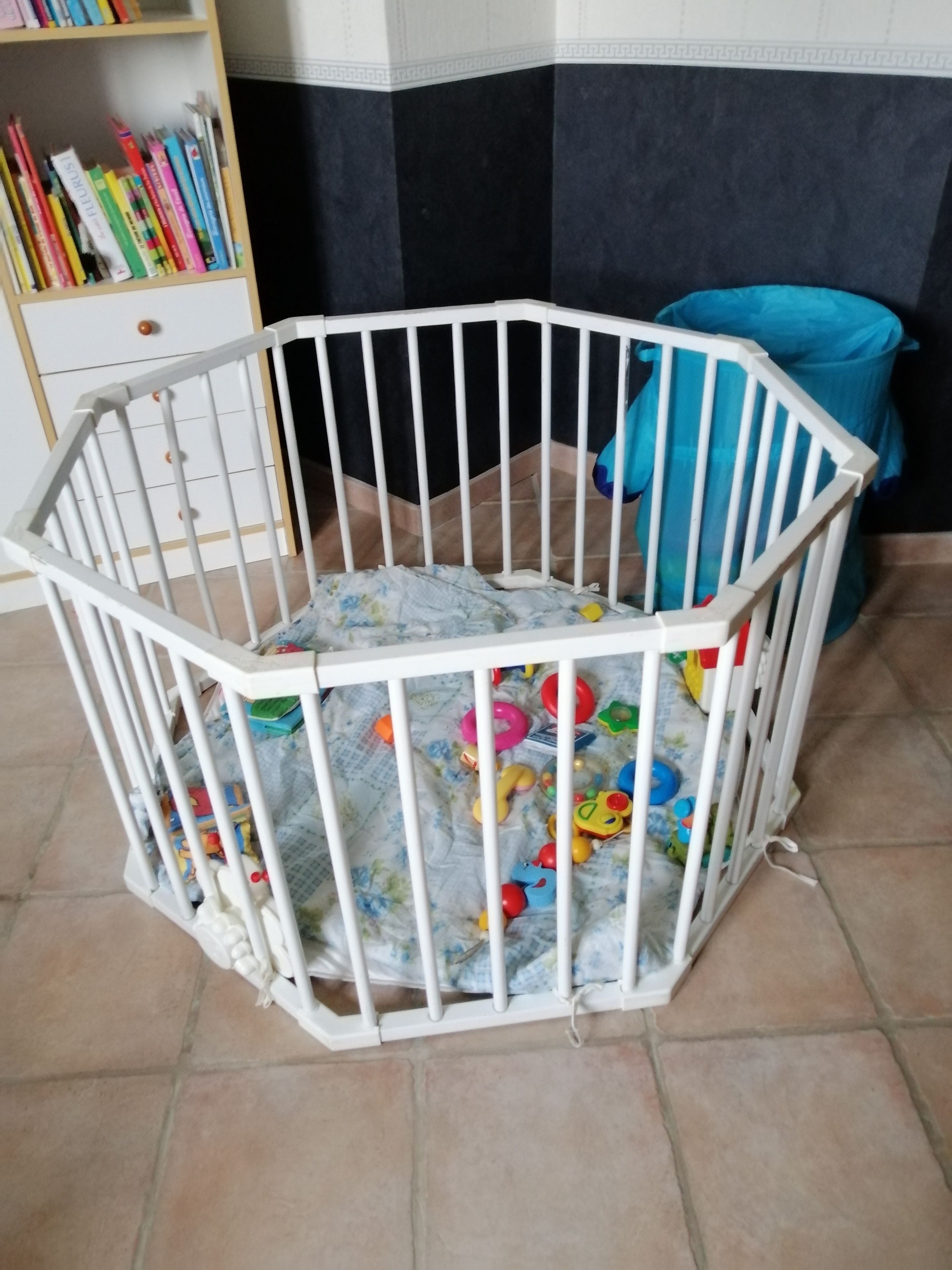 Een wit babyhek met speelgoed erin, een boekenkast met boeken, en een blauwe stoel op de achtergrond.